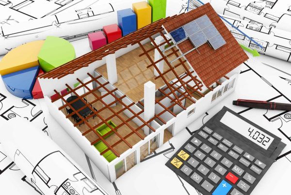 Miniature house with an unfinished roof on building plans next to a calculator, financial charts, and pen.